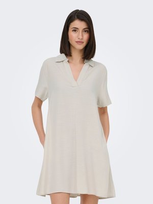 Žena u svijetlo bež haljini s kratkim rukavima i ovratnikom stoji s rukama u džepovima na jednobojnoj svijetloj pozadini.