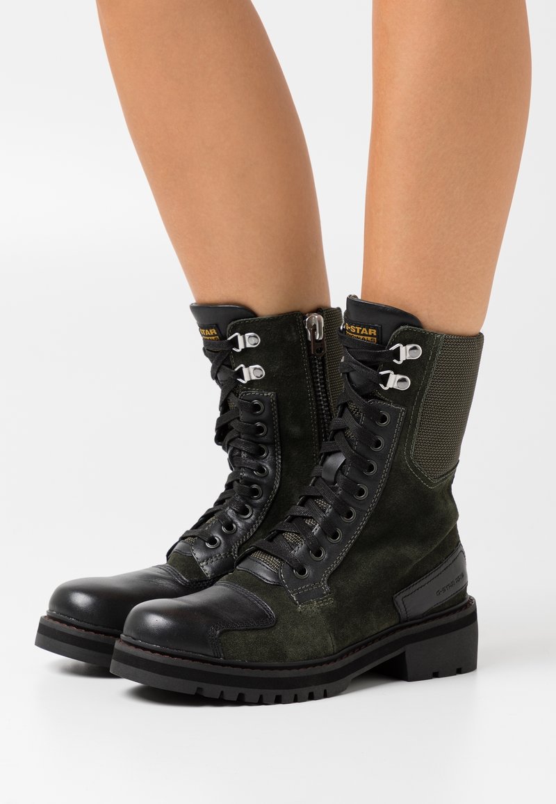 Bottes de combat noires et vert foncé à lacets, avec fermetures éclair latérales, œillets métalliques, semelles épaisses, portées sur jambes nues sur fond blanc.