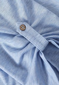 Light blue fabric with a soft texture, featuring a wooden button and a gathered design detail accentuating the neckline.