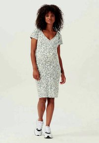 Ziedu raksta pieguļoša kleita gaiši zaļā krāsā ar V veida kakla izgriezumu un īsām piedurknēm, saskaņota ar baltām sporta kedām.