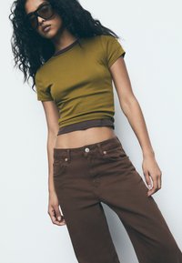 Olijfgroene, aansluitende t-shirt met bruine afwerking, gecombineerd met hoge, bruine broeken met knoopsluiting en zijzakken.