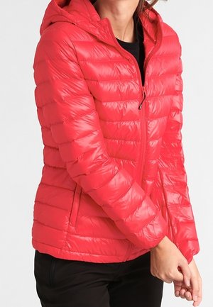 Vrouw draagt een felrode gewatteerde jas met capuchon, dichtgeritst, staand met handen voor zich geklemd.
