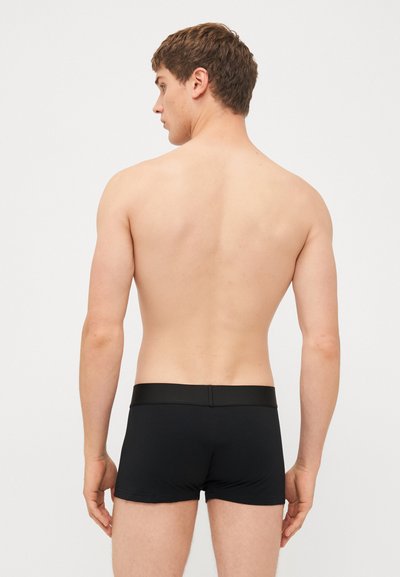 Svarte trunk underbukser med en glatt tekstur, elastisk midjebånd og kort ben-designer. Båret av en mannlig modell som vender bort fra kameraet.