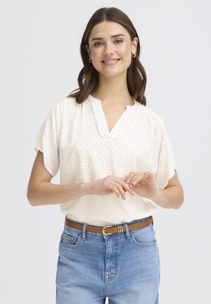 Žena s hnedými vlasmi, oblečená v bielej blúzke s krátkym rukávom a ružovými bodkami, modrých džínsoch a hnedom opasku, usmievajúca sa so založenými rukami.