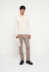 Crème lange mouwen polo shirt, beige broek en zwarte instappers. De persoon houdt een lichtgrijze pet vast. Eenvoudig ontwerp zonder zichtbare patronen.