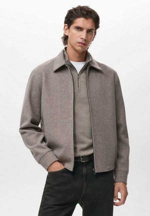 Chaqueta gris con cuello y cierre de cremallera, hecha de una tela texturizada, combinada con un suéter gris claro y jeans oscuros.