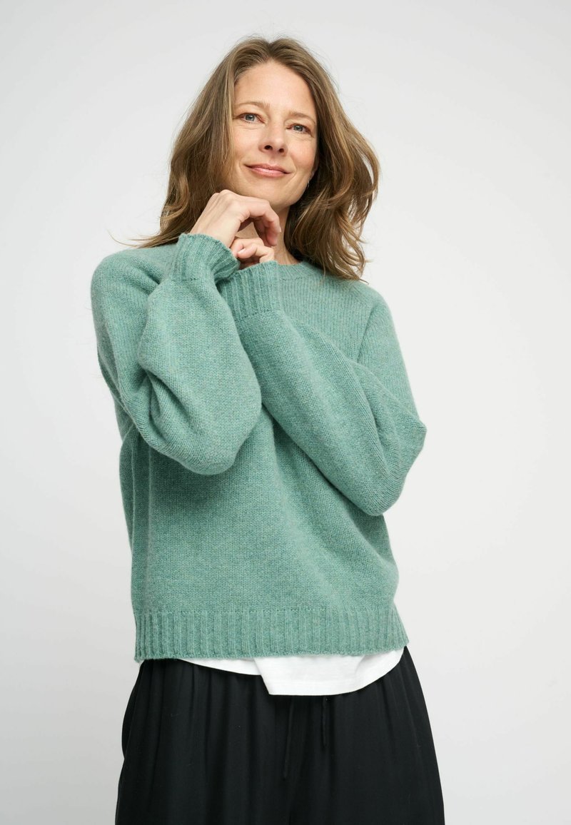 Zachte groene cashmere trui met een ontspannen pasvorm, geribbelde manchetten en zoom, gelaagd over een wit shirt, gecombineerd met losse zwarte broeken.