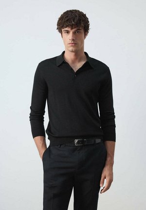 Joven con cabello oscuro, vestido con una camisa polo negra de manga larga y pantalones negros con un cinturón negro, de pie frente a un fondo claro y liso.