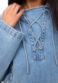 Pullover in denim blu chiaro con scollatura alla marinara, dotato di lacci grigi e occhielli in argento. Dettaglio di cucitura visibile sulle maniche.