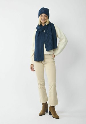 Donkerblauwe gebreide muts en bijpassende sjaal, beide met een geribbelde textuur. Model draagt een crèmekleurige trui en beige flared broek met bruine haklaarzen.