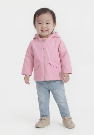 Tout-petit souriant, portant une veste à capuche rose, un jean bleu clair et des chaussures beige, debout devant un fond blanc uni.