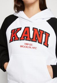 Hanorac alb și negru cu buzunar frontal. "KANI" scris cu litere roșii îngroșate, iar "1989 EST. BROOKLYN, NYC" brodat dedesubt în roșu.