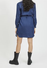 Džínová košilová šaty v tmavě modré barvě, dlouhé rukávy s manžetami na knoflíky, pas s páskem a boční rozparky na spodním lemu. V kombinaci s černými vysokými botami po kolena.