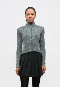 Pelēka faktūras adīta jaka ar augstu apkakli un priek�šējā rāvējslēdzēju, kopā ar plisētu melnu svārku, attēlota uz balta fona.