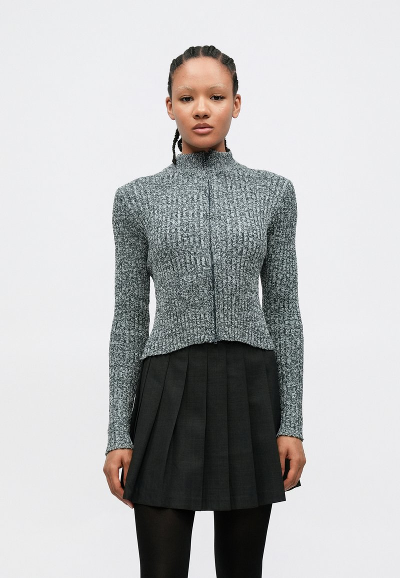 Pelēka faktūras adīta jaka ar augstu apkakli un priekšējā rāvējslēdzēju, kopā ar plisētu melnu svārku, attēlota uz balta fona.