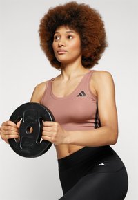 Svart vektskive holdt i hendene; modell iført en rosa sportsbh med svarte striper og høyt liv svarte leggins. Jevn tekstur, atletisk passform.