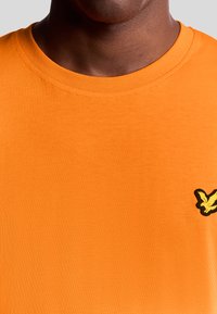 T-shirt di cotone arancione con colletto rotondo, caratterizzata da un logo di uccello ricamato giallo e nero sul lato sinistro. Texture liscia.