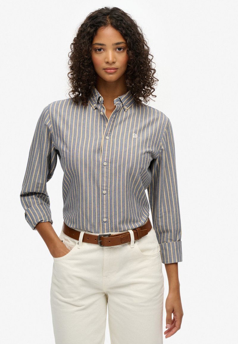 Koszula w paski, zapinana na guziki, w kolorze niebieskim i beżowym, z bawełnianego materiału. Posiada kołnierz, długie rękawy oraz małe logo na piersi, zestawiona z beżowymi spodniami.