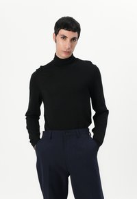 Sort turtleneck sweater lavet af et blødt strikket stof, med lange ærmer. Kombineret med marineblå bukser med en enkel frontknapdetalje.