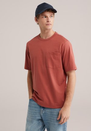 Jaunas vyras dėvi rūdžių spalvos trumpomis rankovėmis kišeninį marškinėlį, šviesiai mėlynas džinsus ir tamsiai mėlyną beisbolo kepurę, žiūri į šoną.