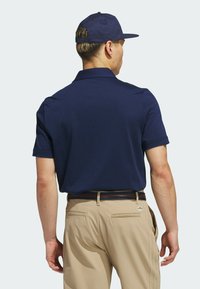 Mørkeblå polo med korte ærmer, laget av pustende stoff. Kombinert med beige bukser og en mørk caps. Synlig logo på kragen.