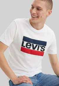 Joven sonriendo, vestido con una camiseta blanca de Levi's con rayas azules y rojas, jeans azules, sentado contra un fondo gris claro liso.
