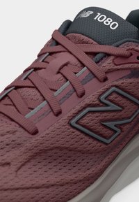 Κοντινό πλάνο ενός μπορντό παπουτσιού για τρέξιμο New Balance 1080 με υφή από πλέγμα, κορδόνια στο ίδιο χρώμα, αντανακλαστικές λωρίδες και μαύρο λογότυπο στο πλάι.