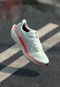 Egy fehér sportcipő rózsaszín részletekkel látszólag lebeg egy szürke felület felett, kiemelve sima dizájnját és hálós textúráját.