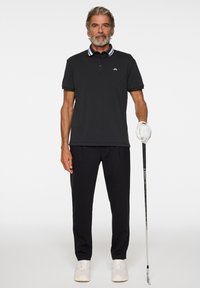 Musta poolopaita, jossa on valkoiset ja laivastonsiniset raidalliset kaulukset, yhdistettynä mustiin housuihin ja valkoisiin tennareihin. Mies pitää golfmailaa kädessään, jossa on golfkäsine.