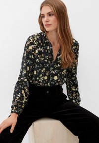 Schwarzblumige Bluse mit grünen, gelben und lila Blumen, langen Ärmeln, Knopfleiste und kombiniert mit schwarzen Cordhosen.