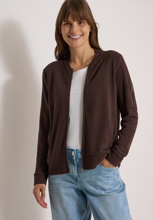 Femme souriante portant une veste zippée marron foncé avec une poche zippée sur la manche, une chemise blanche et un jean bleu clair, debout devant un fond uni.