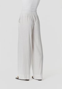 Inimene, kes kannab lahtisi valgeid laiade jalgadega pükse ja musti sandaale, seisab ühevärvilise valge tausta ees.