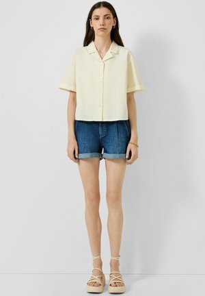 Femme portant une chemise jaune pâle à manches courtes boutonnée, un short en jean retroussé et des sandales à plateforme beiges à brides, debout devant un fond uni.