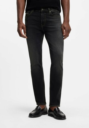 Unterkörper einer Person, die schwarze, ausgewaschene Slim-Fit-Jeans mit Taschen und schwarze Leder-Loafer auf schlichtem Hintergrund trägt.