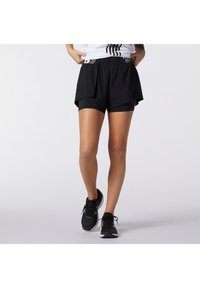 Pantalones cortos deportivos negros con una capa de malla, una cintura elástica y pantalones cortos de compresión negros integrados por debajo. Se llevan zapatillas de correr negras.