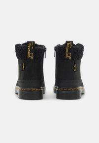 Cizme negre până la gleznă cu un material superior moale și texturat, guler din blană de miel și fermoare. Prezintă o talpă groasă, cu striații și accente de cusătură galbenă.