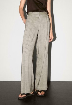 Personne portant un pantalon à carreaux taille haute à jambes larges, un haut noir sans manches, des sandales noires de type tongs, debout avec une main dans la poche.