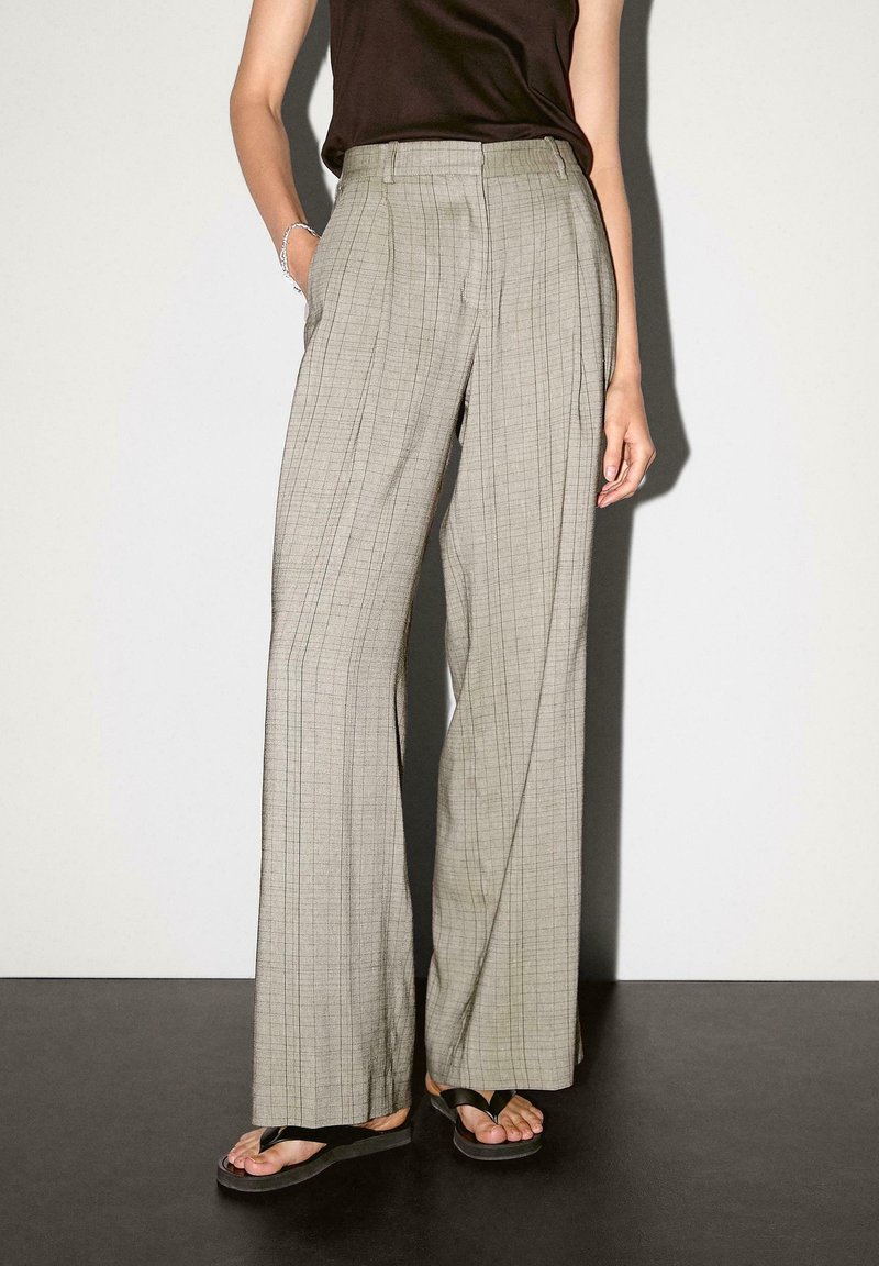 Personne portant un pantalon à carreaux taille haute à jambes larges, un haut noir sans manches, des sandales noires de type tongs, debout avec une main dans la poche.