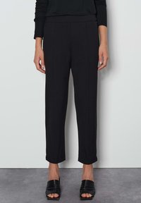 Zwarte broek met een gladde textuur, taps toelopend design en enkellange snit. Heeft subtiele voornaden en een elastische tailleband.