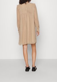 Beiges Polka-Dot-Kleid mit rüschierten Schulternähten, runder Halslinie und Rückenschlitzdetail. Durchsichtiger, leichter Stoff; lange Ärmel. Schwarze Schuhe.