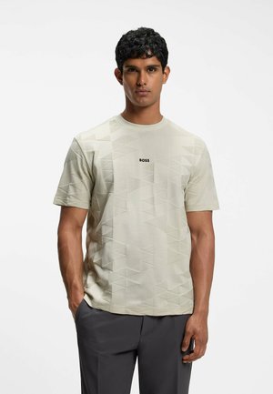 Mann mit kurzen lockigen Haaren, der ein hellbeiges gemustertes T-Shirt mit dem Text "BOSS" und dunkle Hosen trägt, steht mit einer Hand in der Tasche.