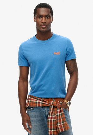 Camiseta de algodón azul con cuello redondo y mangas cortas, que presenta un logo naranja en el pecho. Se lleva con una tela de cuadros atada a la cintura.