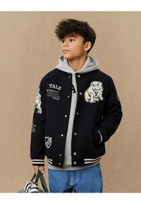 Námornícky modrá varsity bunda s vyšitým logom Yale, grafikou buldoga a bielymi pruhmi. Nosená cez šedú mikinu s kapucňou, spolu s džínsami.