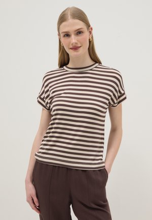 Femme portant un t-shirt à manches courtes rayé horizontalement marron et blanc et un pantalon marron, debout devant un fond clair uni.