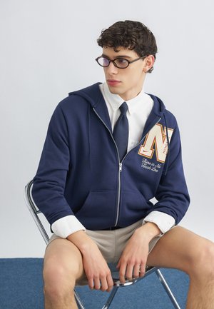 Młody mężczyzna w okularach, granatowej bluzie na zamek, białej koszuli i granatowym krawacie, siedzący na metalowym krześle na jednolitym tle.