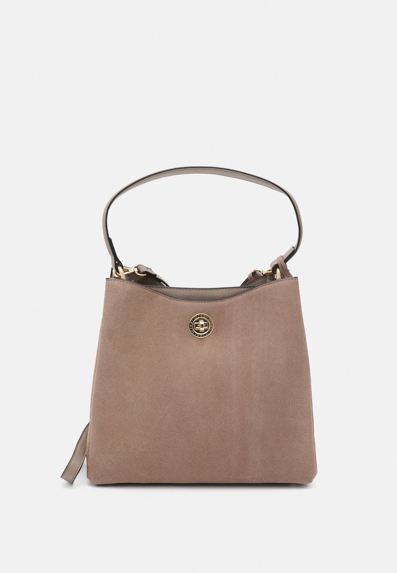 Sac à main en daim taupe avec une texture lisse, une poignée supérieure et un détail en métal doré circulaire à l'avant. Design simple et moderne.