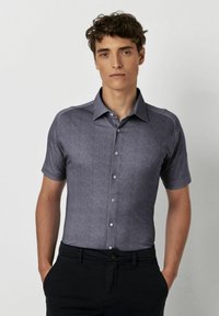 Kurzärmliges, knöpfbares Hemd aus dunkelblauem Stoff mit einem strukturierten Muster und einem Kragen; getragen mit schwarzen Hosen.