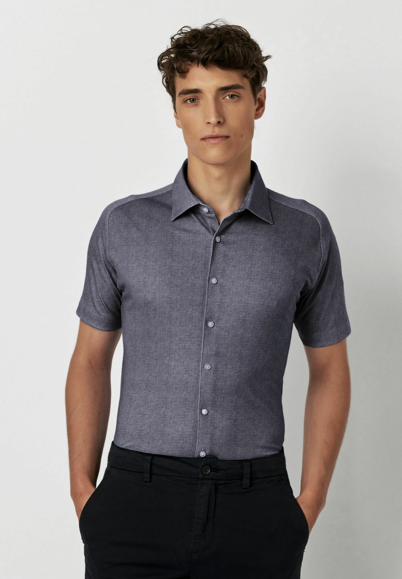 Kurzärmliges, knöpfbares Hemd aus dunkelblauem Stoff mit einem strukturierten Muster und einem Kragen; getragen mit schwarzen Hosen.