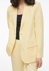Žena u blijedožutom odijelu s dvorednim sakoom i hlačama u istoj boji, uparenom s crnim topom ispod.