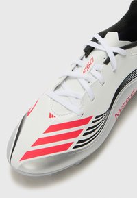 Witte voetbalschoen met rode en zwarte strepen, voorzien van een glad bovenwerk van synthetisch materiaal, witte veters en zilverkleurige details op de zool.
